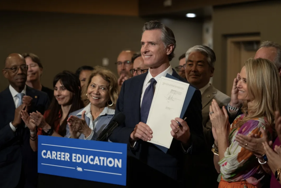 Governor Newsom smiles and holds up a piece of legislature at a podium in a press conference.