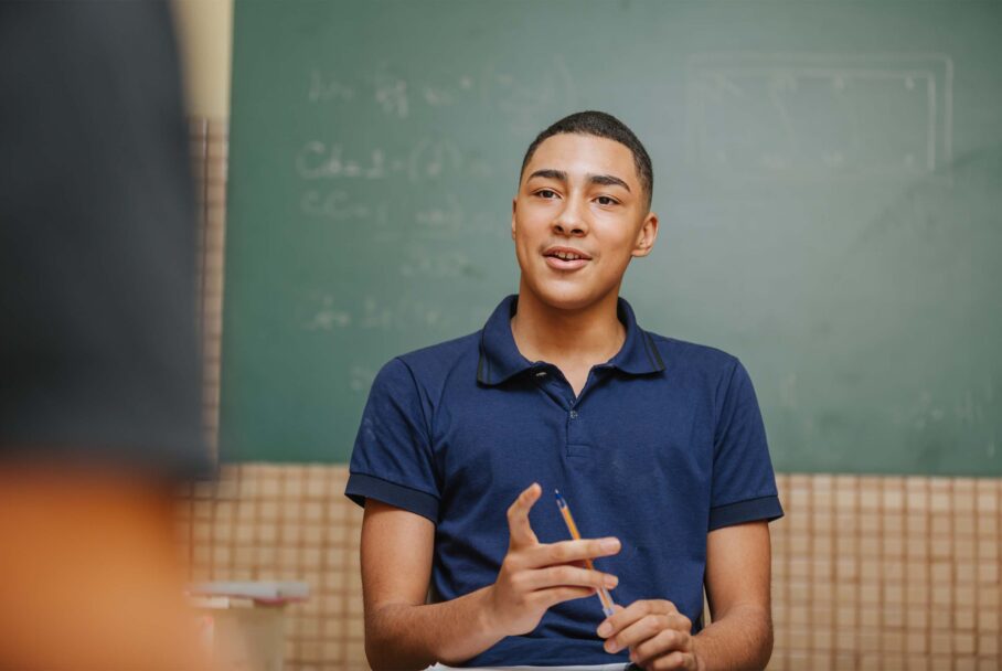 Male student holds a pen in front of a chalkboard and talks to someone out of frame of the image.