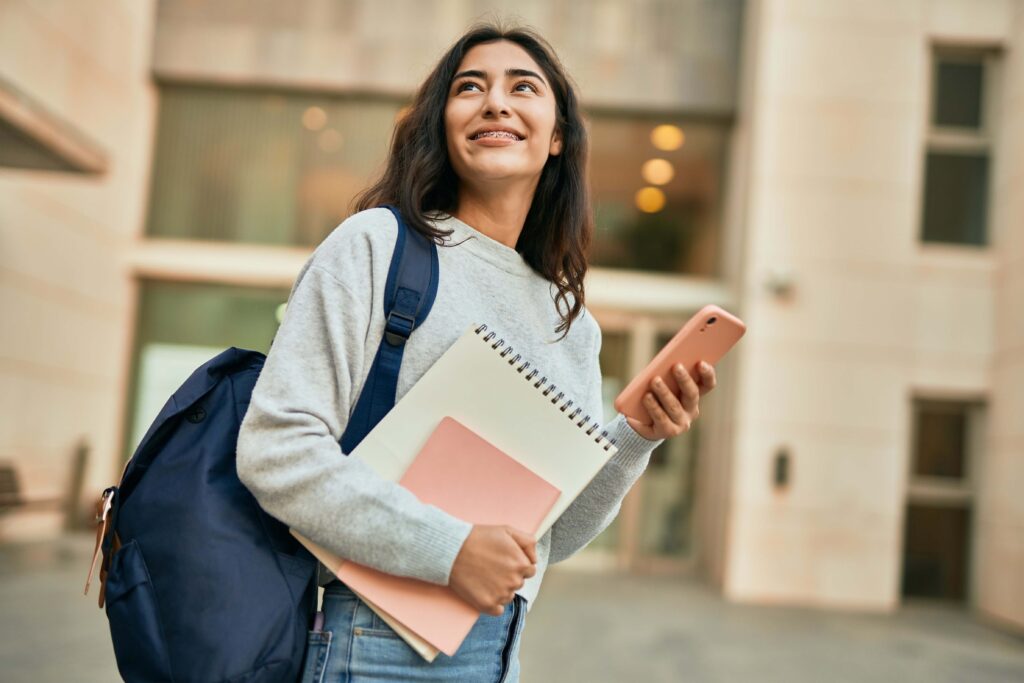 Girl with a backpack on her shoulder, holding a notepad and her phone smiles and looks upwards.