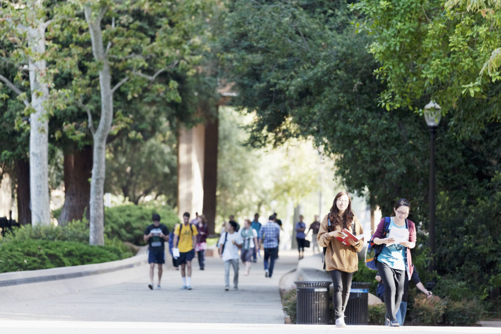 The location is University of California, Los Angeles. Two Asian female students are walking in the foreground. A larger group of students are walking in the background.