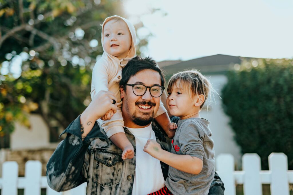 Smiling father holding a small child on his hip and a toddler on his shoulders. 